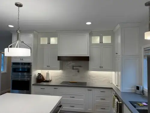 White kitchen with double wall ovens, glass-front upper cabinets, stone backsplash and wood range hood